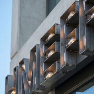 A daytime photograph of industrial-style, open-face metal channel lettering with exposed bulbs mounted on a concrete building facade.