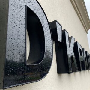 Exterior dimensional signage made of black acrylic and metal shown close-up with rain droplets on the surface during overcast weather.