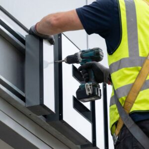 A close-up photograph of a technician's gloved hands using a tool to install a large dimensional sign component onto a building exterior.